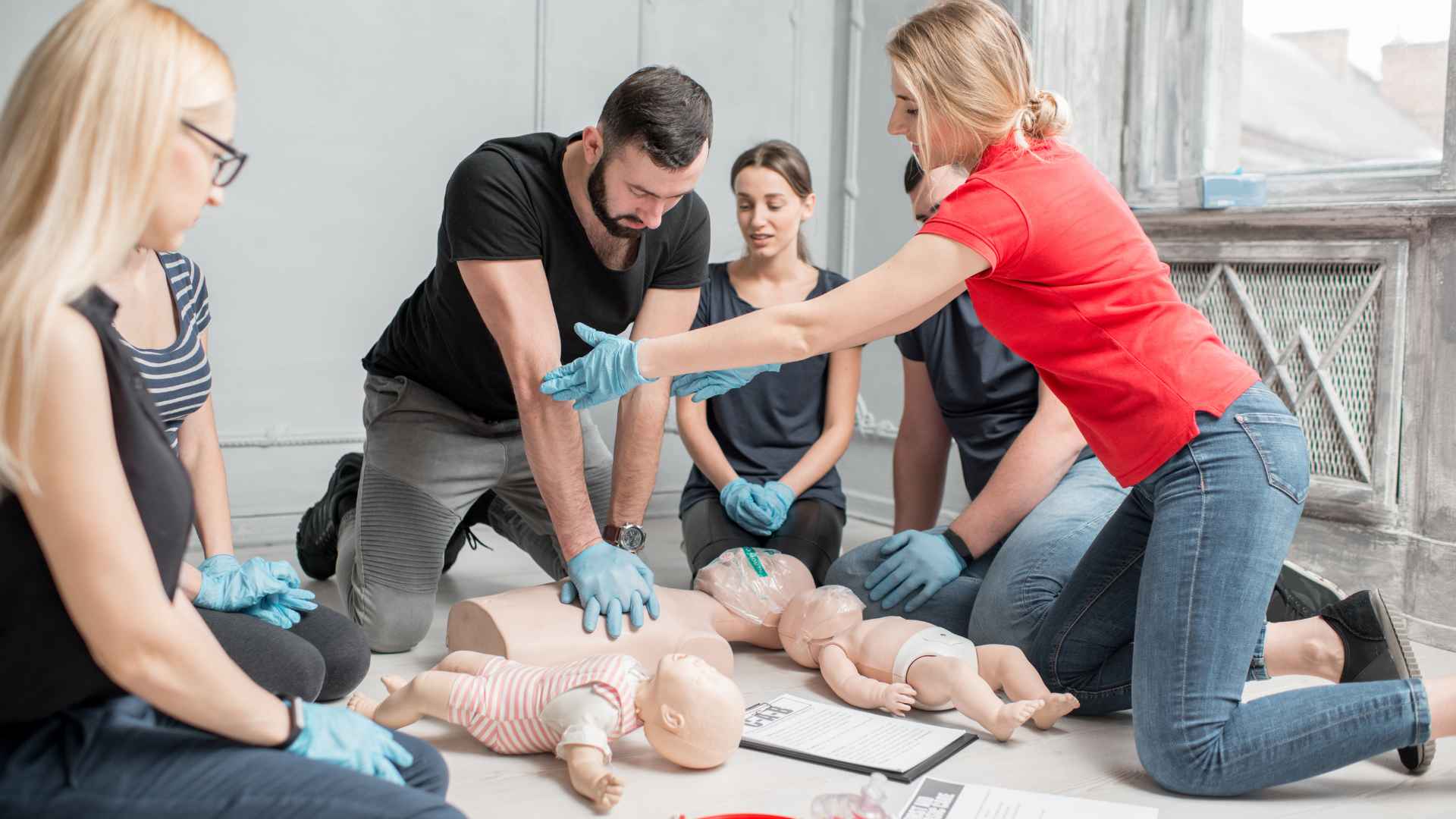 a first aid and cpr training class of men and women. a man is practicing cpr on a manikin and a female instructor is guiding him