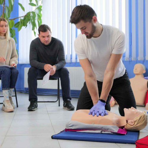 man practicing cpr on a manikin with additional students sitting in the background on chairs