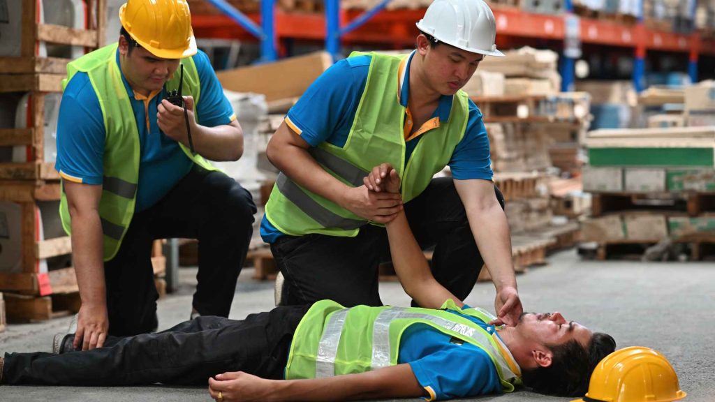warehouse worker laying on the ground unconscious. his yellow hard hat fell off. another worker is checking on him and a third worker is using a radio