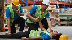 warehouse worker laying on the ground unconscious. his yellow hard hat fell off. another worker is checking on him and a third worker is using a radio