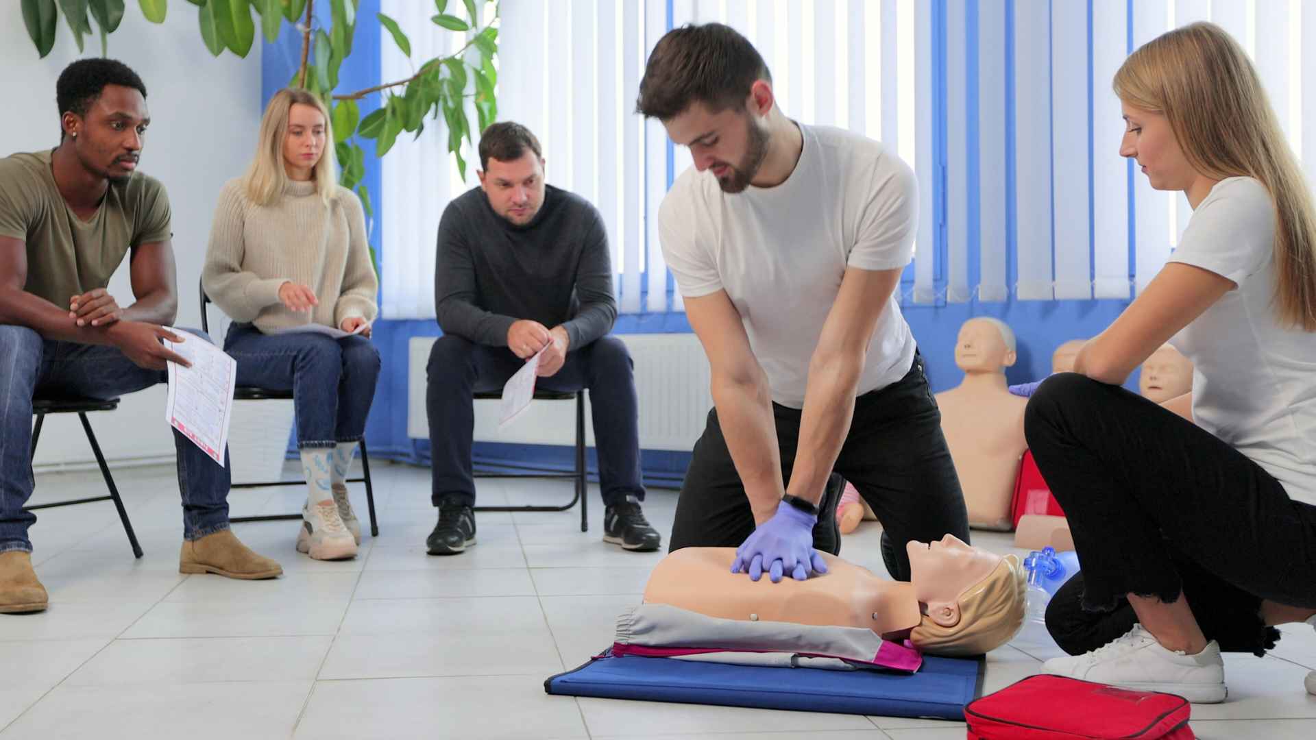 man practicing cpr on a manikin with additional students sitting in the background on chairs