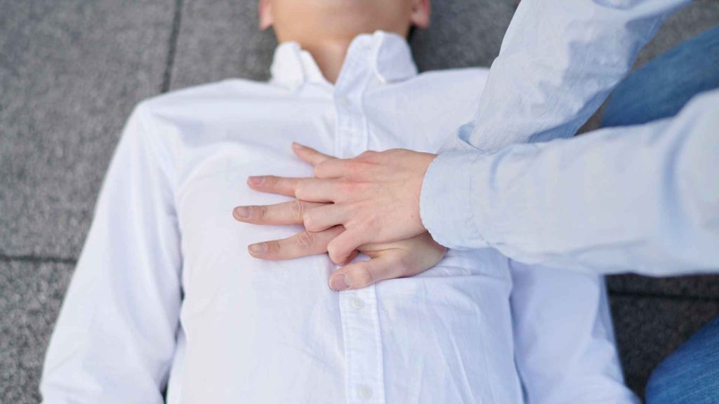 close up of someone performing chest compressions on a man laying down on the ground in a white dress shirt