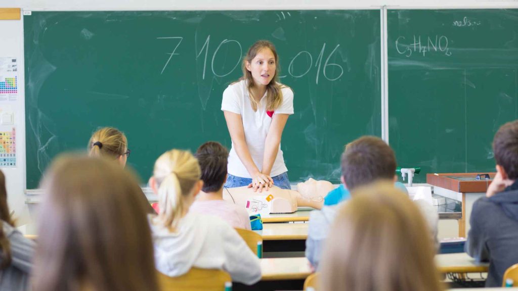 a teacher displaying how to perform cpr on a manikin in front of a of teenagers