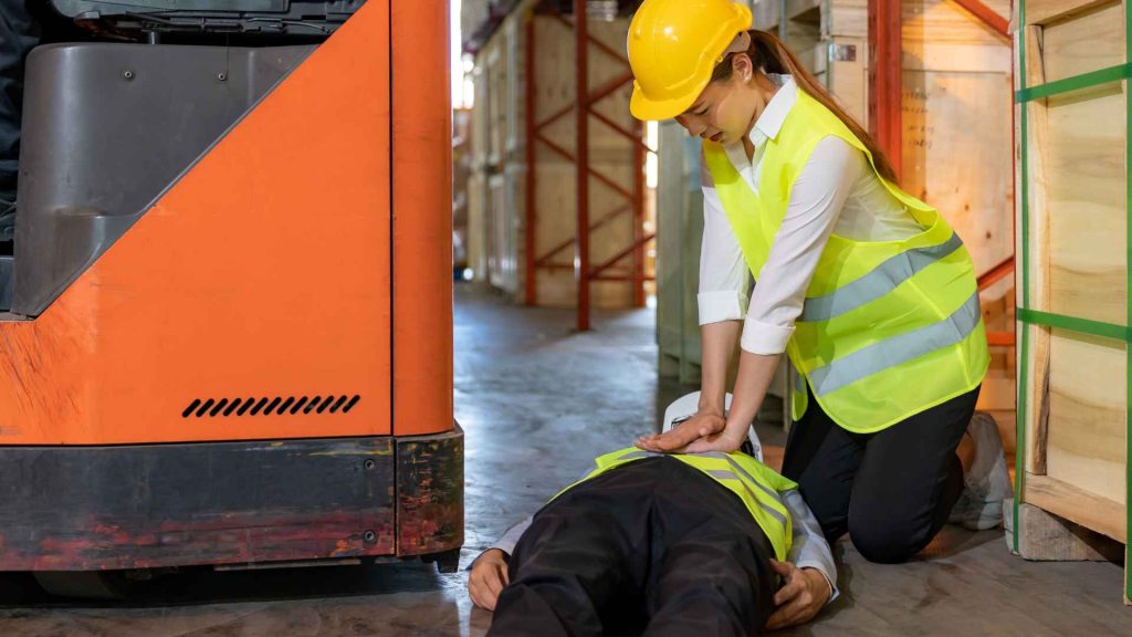 woman wearing safety vest and yellow hard hat performing cpr on another worker. they are in a warehouse.