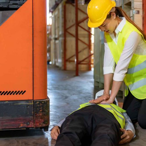 woman wearing safety vest and yellow hard hat performing cpr on another worker. they are in a warehouse.