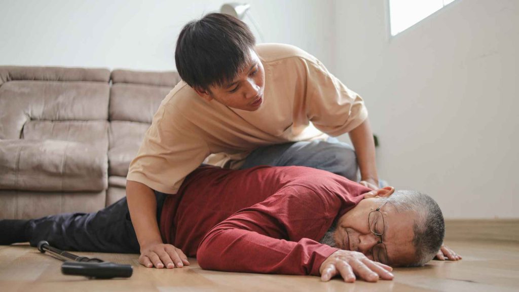 older man laying face down on the floor who may have suffered from cardiac arrest. there is a younger man leaning over him and checking if he is ok.