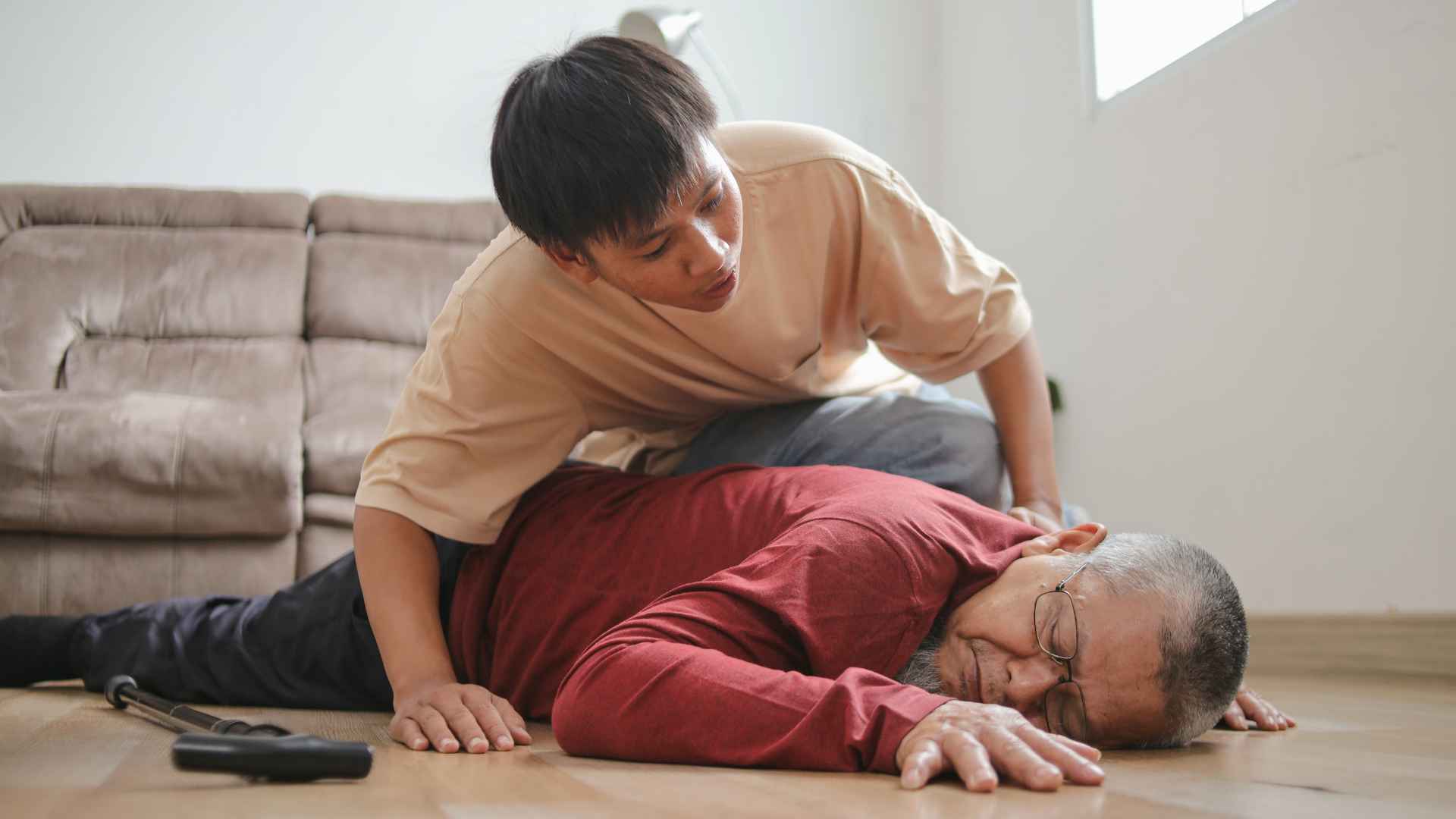 older man laying face down on the floor who may have suffered from cardiac arrest. there is a younger man leaning over him and checking if he is ok.