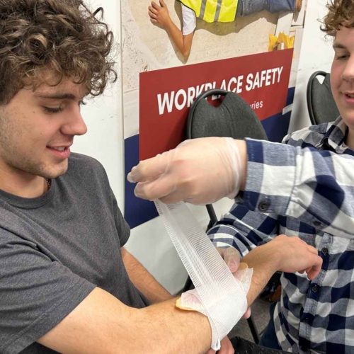 two male students in a first aid training room practicing life-saving skills. the male on the right is practicing applying a bandage to the male on the left's arm.