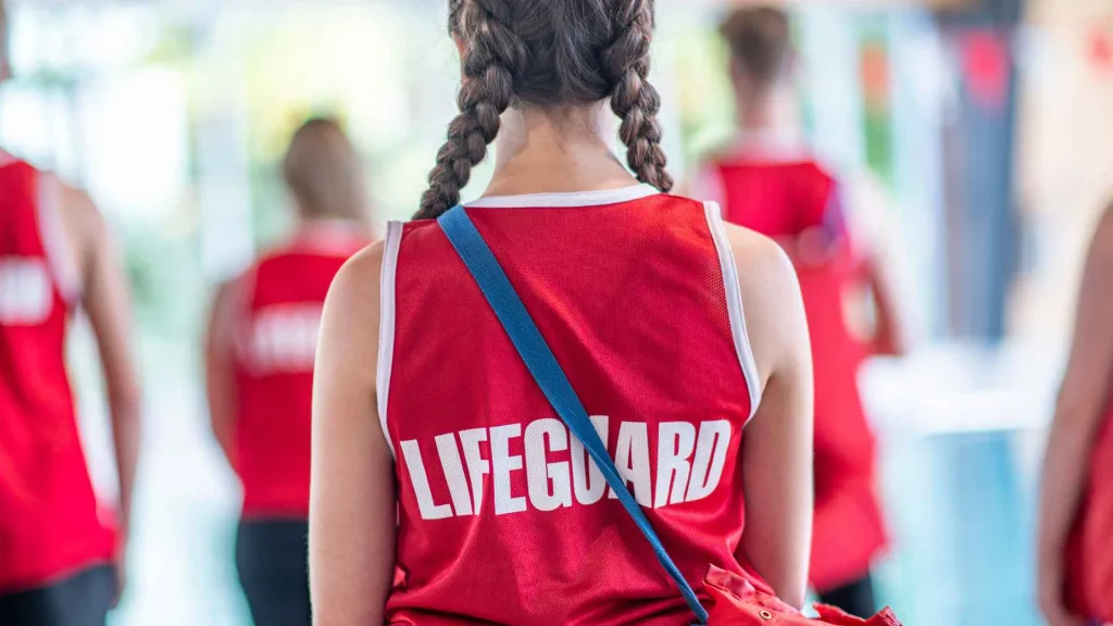 Girl with two braids wearing a red lifeguard shirt facing her back to the camera. There are more lifeguards standing in front of her. They are training to be lifeguards.
