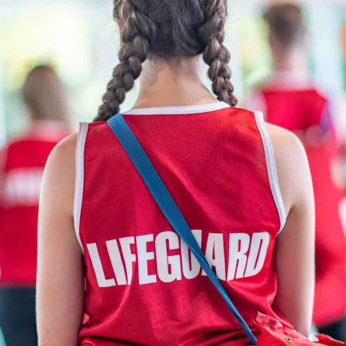 Girl with two braids wearing a red lifeguard shirt facing her back to the camera. There are more lifeguards standing in front of her. They are training to be lifeguards.